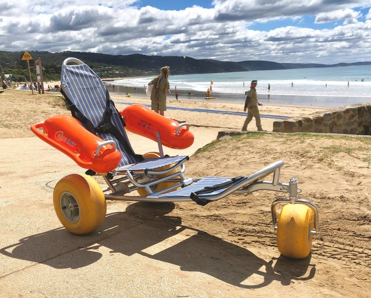 A Mobi-Chair Floating Beach Wheelchair sits on the pavement with the Lorne accessible beach in the background. There are some people walking towards the beach on the pavement. 
