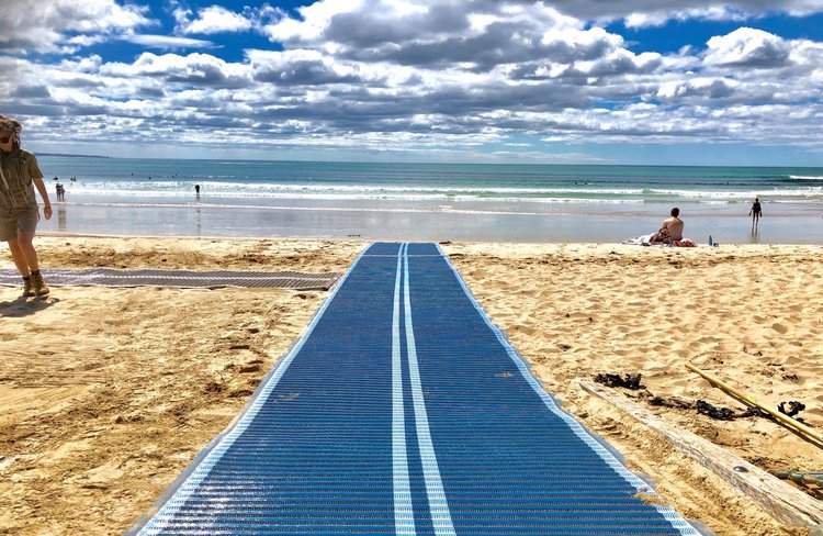 A photo of beach access matting leading over soft sand and towards the ocean. There are people on the beach, some sitting and some standing.
